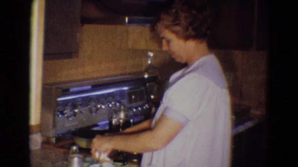 In the 1969 kitchen scene, the woman looks down at the stovetop while continuing breakfast preparation near a pan and eggs.
