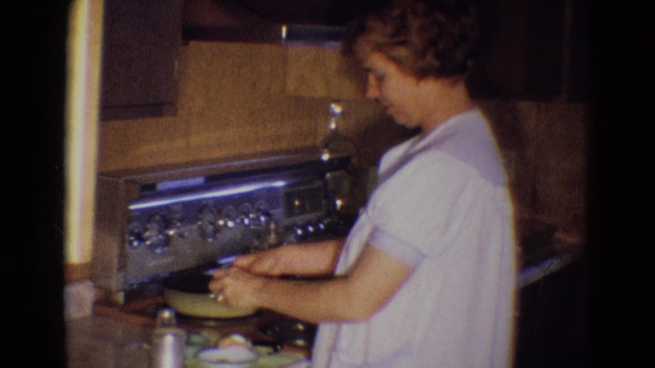 Woman in a pale dress stands at a kitchen stove working over a skillet.