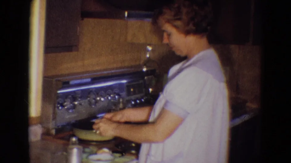 In Diller, Nebraska, 1969, a woman in a light dress cooks at a stovetop beside dark cabinets and counter items.