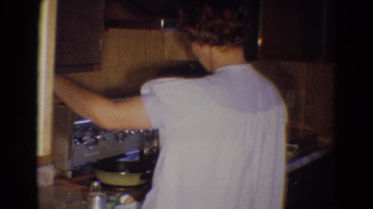 Woman seen from behind reaches toward the stovetop in a small kitchen.