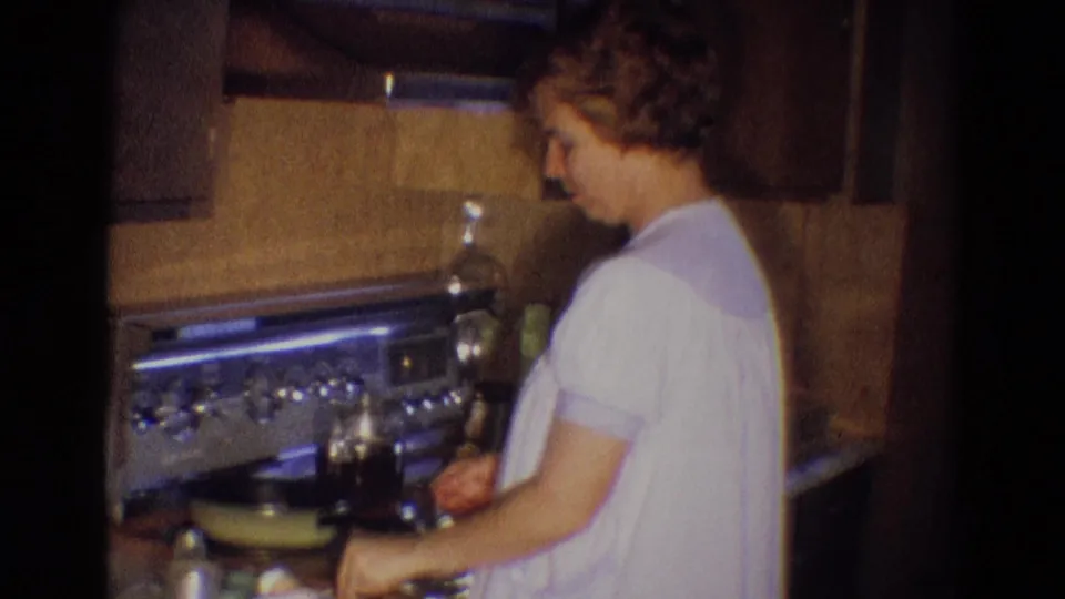 A woman continues cooking at the 1969 Diller kitchen stove, with a skillet, controls, cabinets, and counter objects visible.