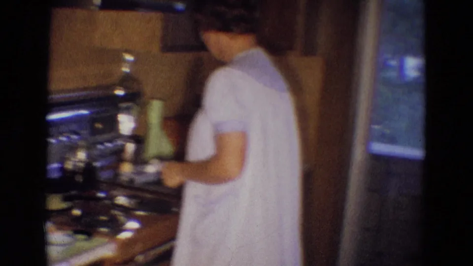 The woman stands beside the stove near a window in the Diller, Nebraska home kitchen, with cookware and counter items visible.