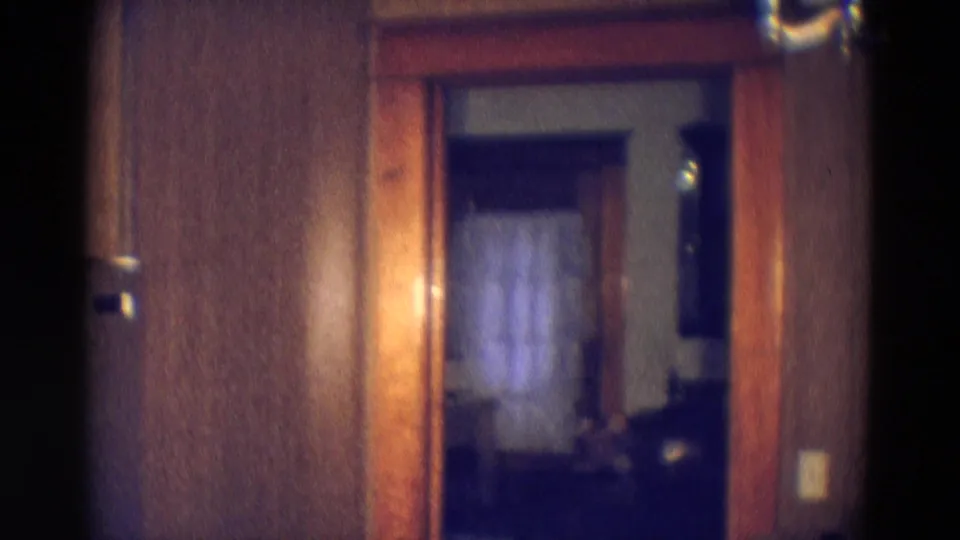 1969 Diller, Nebraska home interior view through a wood-trimmed doorway toward a dark room with curtained window and nearby switch plate.