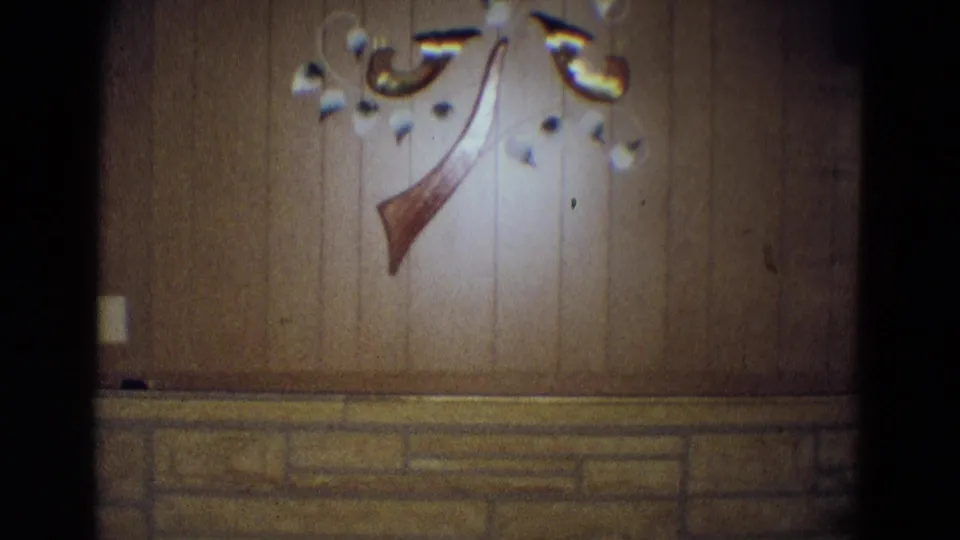 1969 Diller, Nebraska home-movie frame showing a wood-paneled recreation-room wall, stone wainscot, and decorative wall hanging.