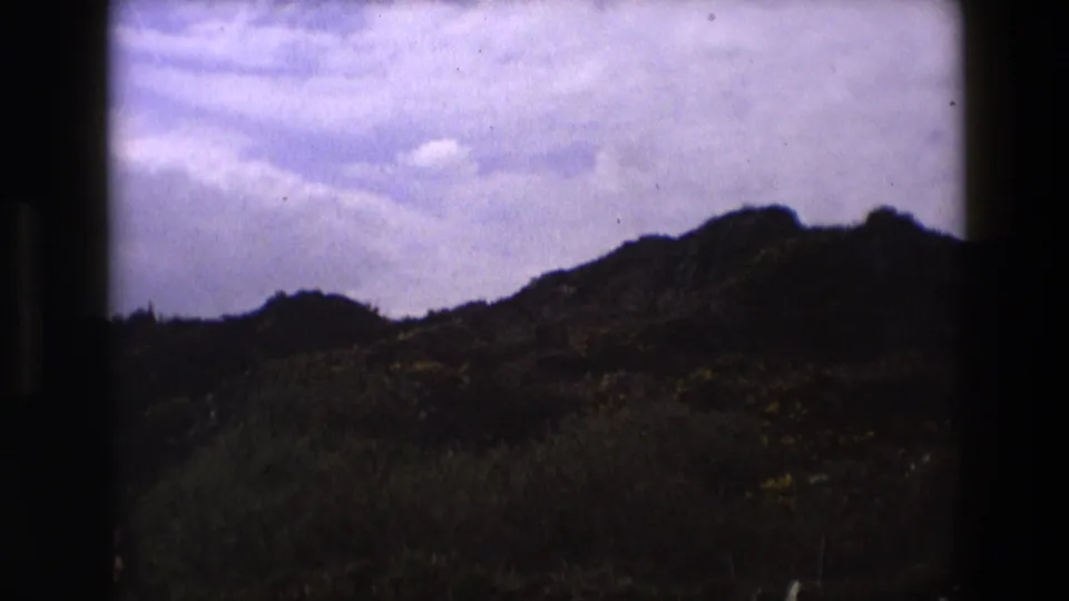1969 England 8mm frame showing a darker ridge or hillside with foreground grasses and a partly cloudy sky.