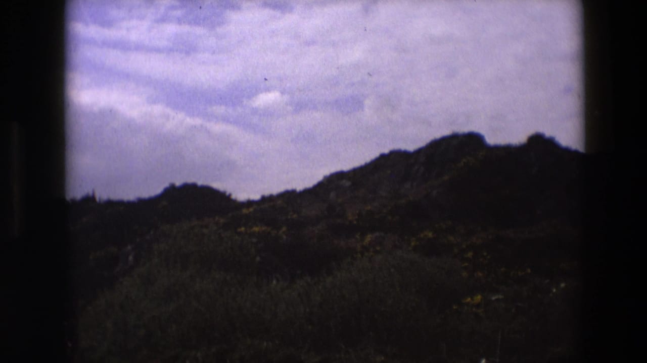 Dark low hillside beneath a pale partly cloudy sky, framed by 8mm film borders.