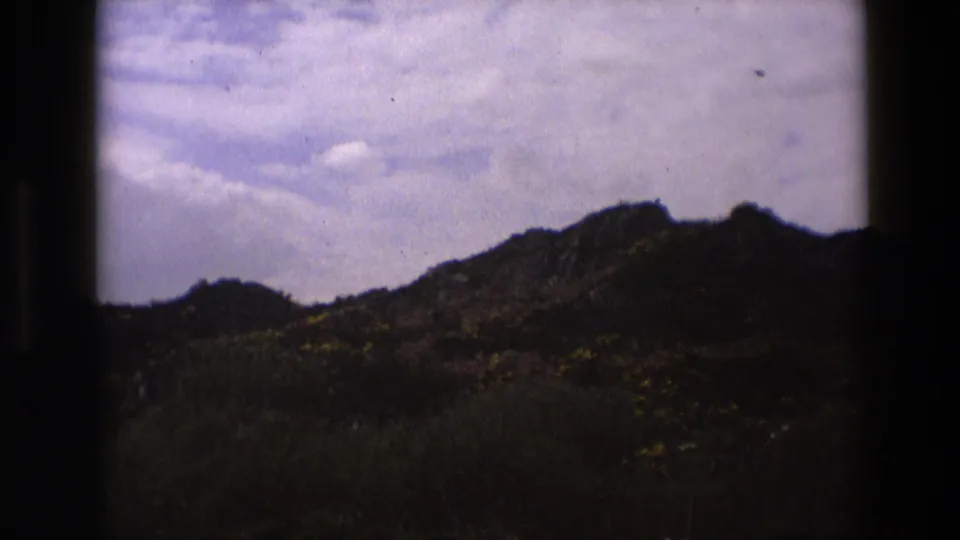 1969 England 8mm frame showing a rugged, shrub-covered hillside with dark foreground vegetation and a lightly clouded sky above.