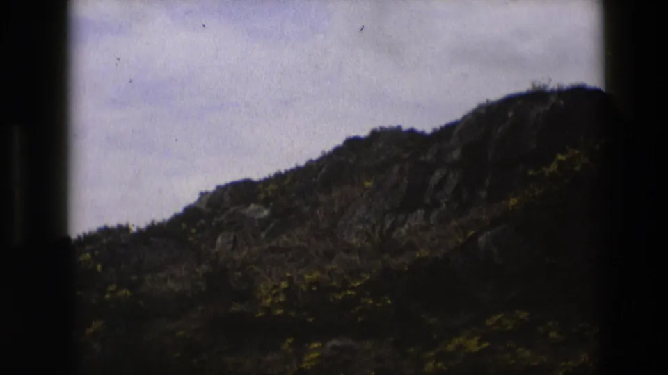 1969 England 8mm frame of a dark rocky hillside with brush, yellow wildflowers and a cloudy sky.