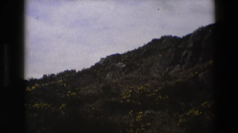 1969 England home-movie frame showing rocky roadside terrain, scrub growth and muted overcast light.