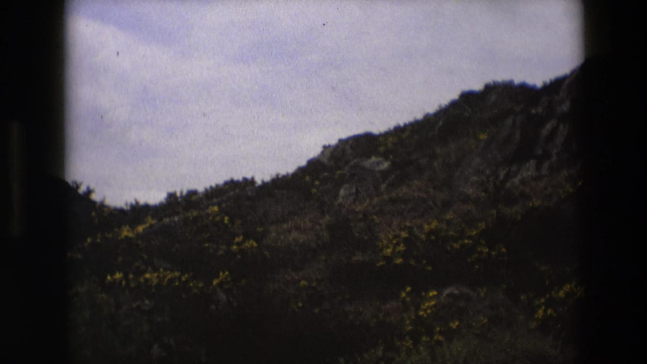 1969 England 8mm frame showing a cloudy sky over a dark, rocky hillside with low scrub and dry vegetation.
