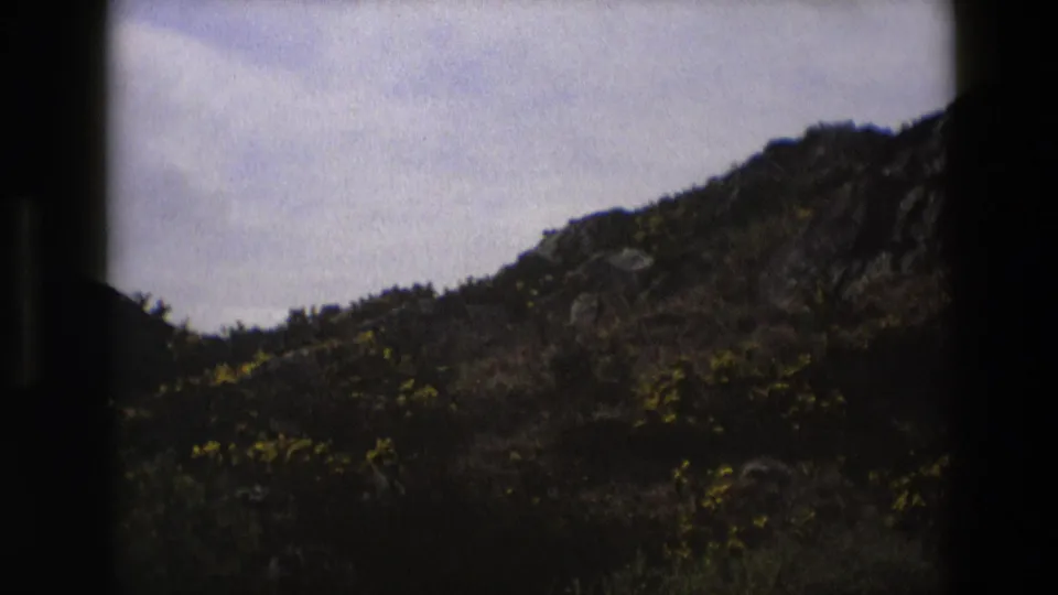1969 England 8mm frame with a pale cloudy sky, rocky slope, and scrubby hillside vegetation.