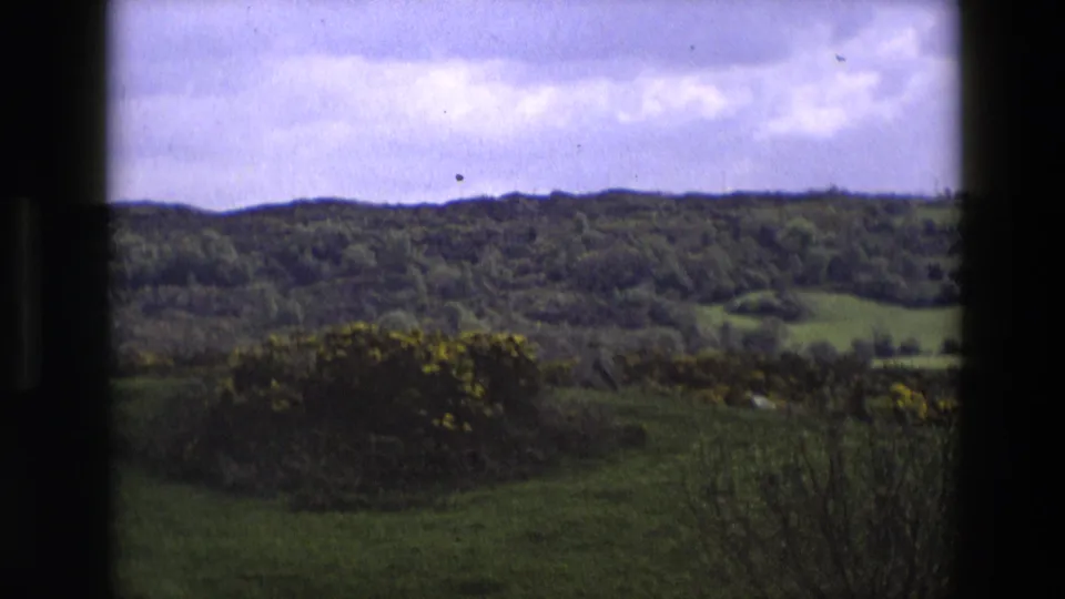 Soft 8mm frame of rural England with distant tree-covered hills, green fields, yellow gorse, cloudy sky, and visible film texture.
