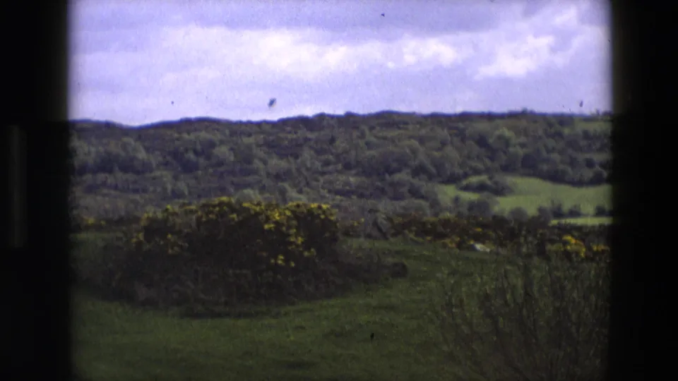 1969 home-movie landscape in England showing rolling hills, woodland, fields, flowering gorse, and dark framing from a window or vehicle edge.