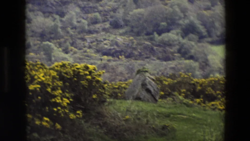 1969 England 8mm frame showing a weathered stone structure in a grassy field near gorse and soft wooded hills.
