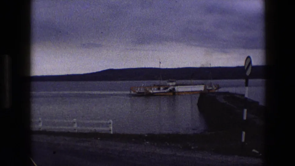 1969 England 8mm frame showing a yellow and white ferry near a stone pier, with calm bay water, low hills, and window-frame edges.