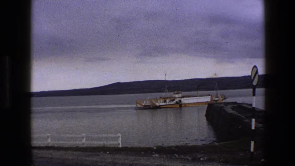 1969 England 8mm frame showing the ferry shifted slightly in view, with white foreground railings, a stone pier, calm water, and heavy clouds.