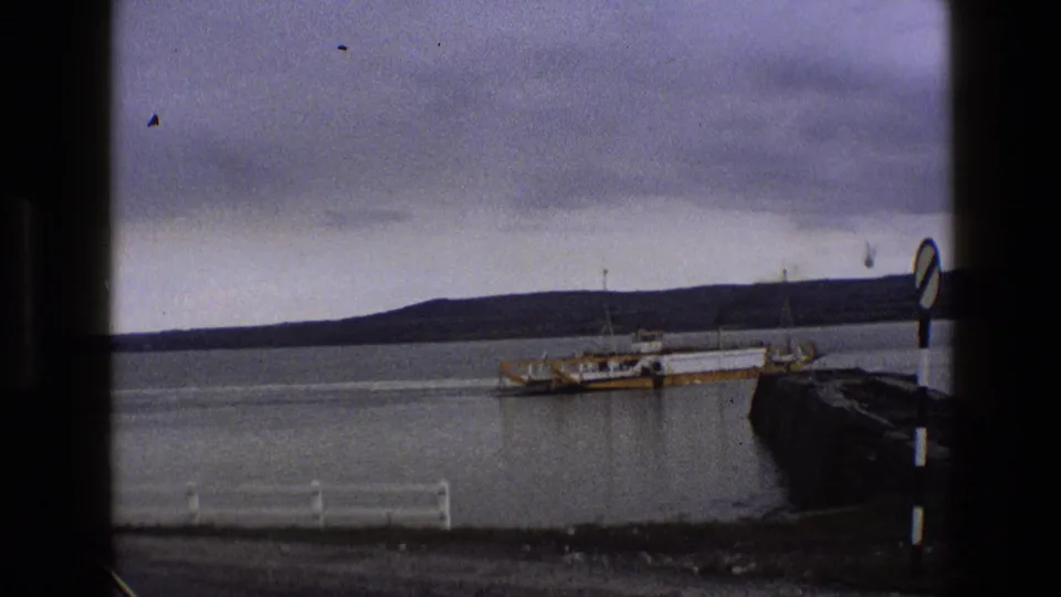 1969 England coastal frame with a yellow and white ferry by a dark pier, a striped sign at right, still water, and low hills in the distance.