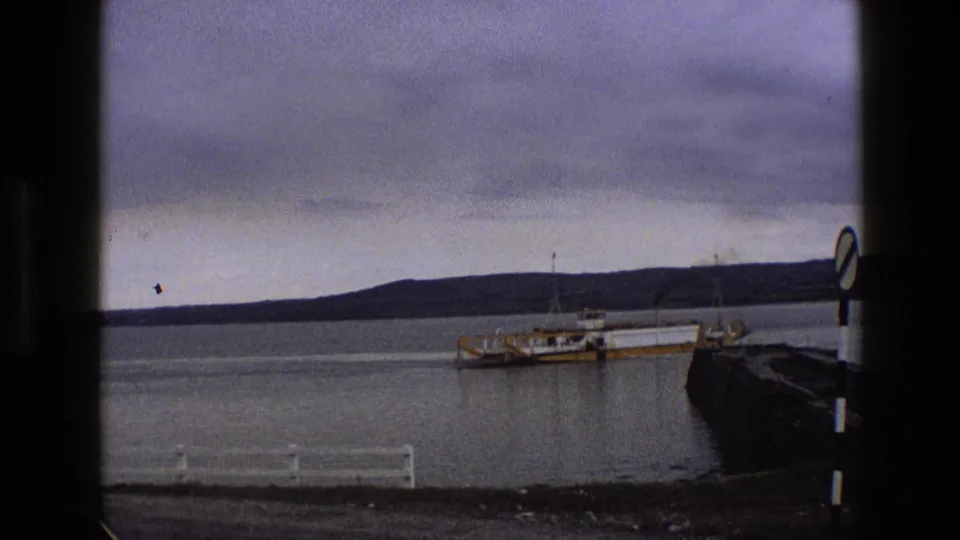 1969 England 8mm frame showing a yellow and white ferry moving across calm harbor water below an overcast sky.
