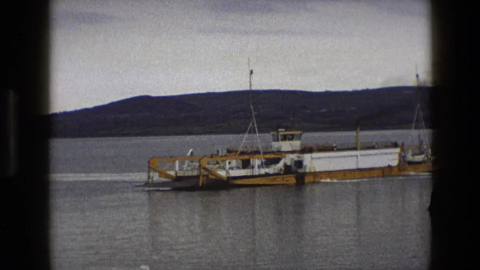1969 England 8mm frame showing the ferry broadside in a quiet harbor, with a muted hillside beyond the water.