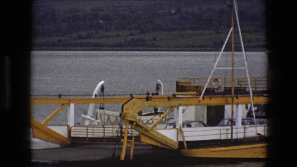 Closer 8mm view of the ferry’s yellow ramp and white deck area, with wide water and a wooded hillside beyond.