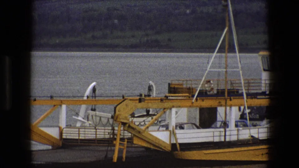 The ferry passes close to camera, showing yellow metalwork, white cabin edges, railings and possible distant people on deck.