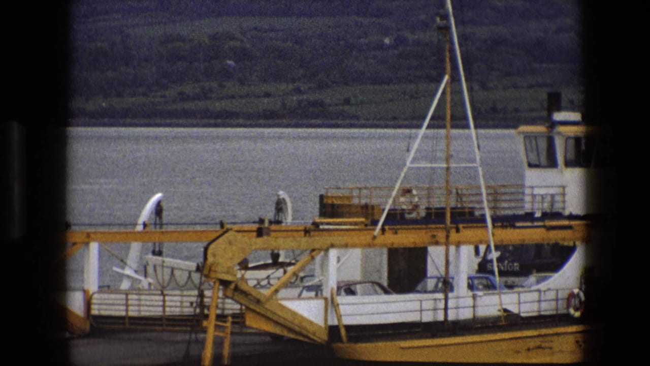 1969 England 8mm frame of a ferry docked beside a yellow gangway, with cars on deck and hills across the water.