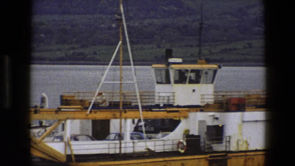 1969 England 8mm frame showing the ferry cabin, open vehicle deck, cars, and a partial vessel marking near the dock.
