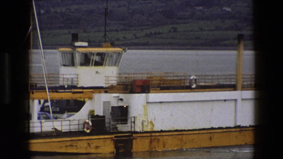 1969 England 8mm frame of the ferry deck and cabin, with railing, lifebuoy, yellow dock structure, and shoreline behind.