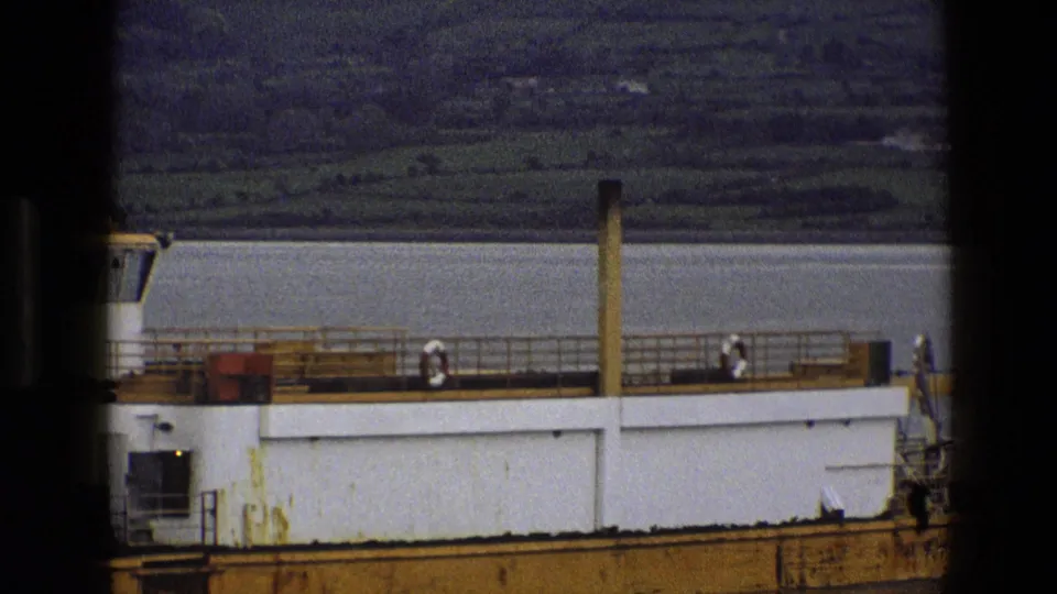 1969 England 8mm frame showing a ferry deck section with railings, lifebuoys, calm water, and green hills beyond.