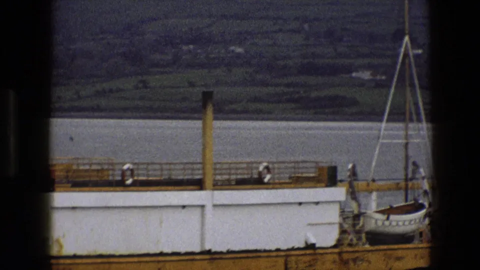 1969 England 8mm frame showing a yellow-hulled vessel with white sides, deck railings, and distant figures, viewed through a dark foreground frame.