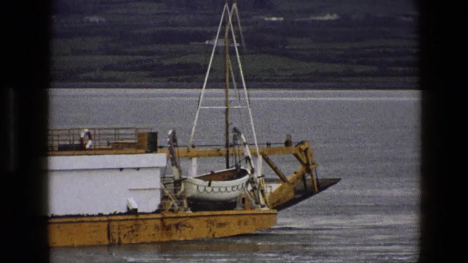 A closer 1969 England home-movie view of the vessel's stern area, with a secured lifeboat, railings, and calm water behind it.