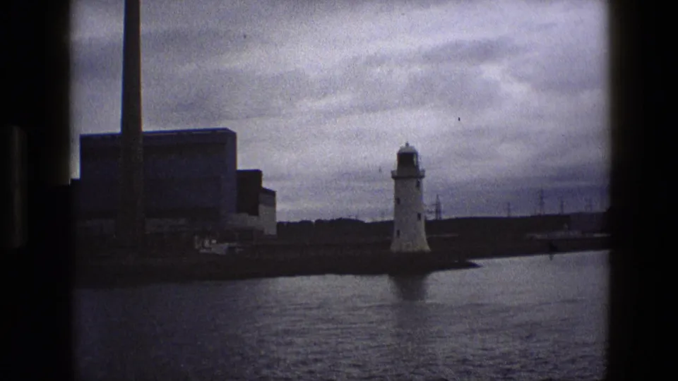 1969 England coastal view with a white lighthouse, dark industrial buildings, a tall chimney, water, and overcast sky.