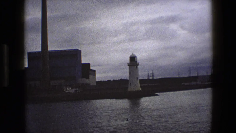 A later 1969 England frame shows the lighthouse and adjacent industrial waterfront across calm water, with muted color and heavy sky.