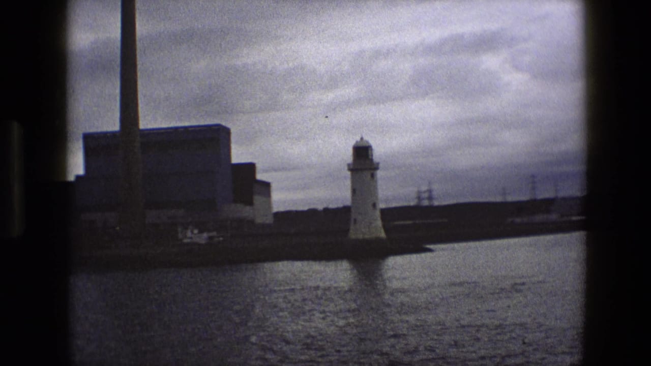 White lighthouse across harbor water with industrial building, chimney, and cloudy sky.