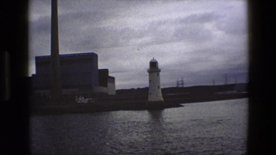 1969 England home-movie frame showing a white lighthouse by water with an industrial building and tall chimney behind it.