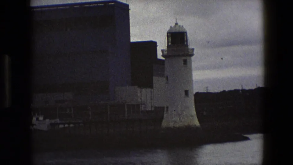 1969 England 8mm frame with a white lighthouse at the harbor edge and a blocky industrial building in the background.
