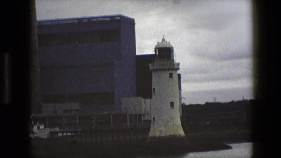 1969 England frame showing the lighthouse near a dockside edge, with water, pilings, and a large industrial building behind.