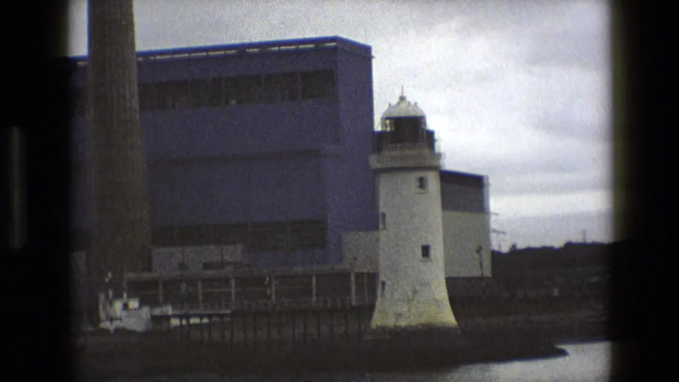 1969 England home-movie frame with the lighthouse beside industrial waterfront buildings and a tall chimney partly visible at left.