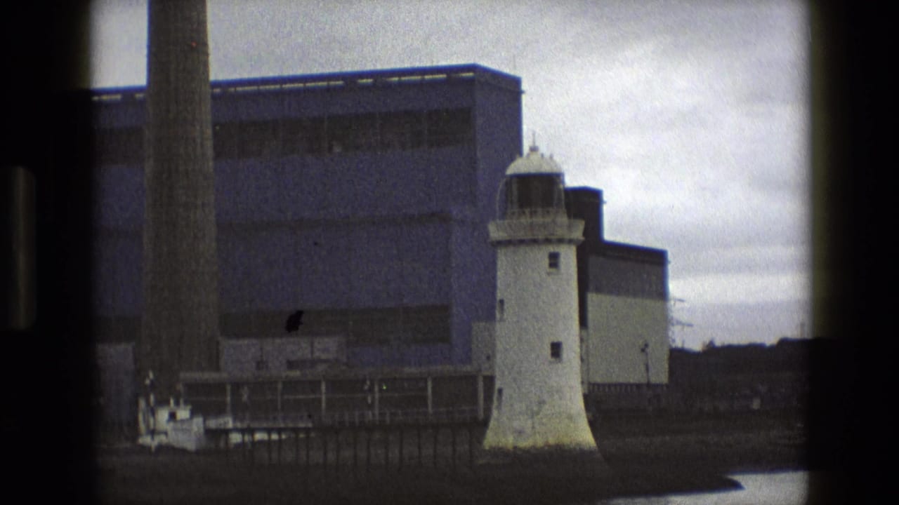 White lighthouse beside an industrial waterfront building and tall chimney.