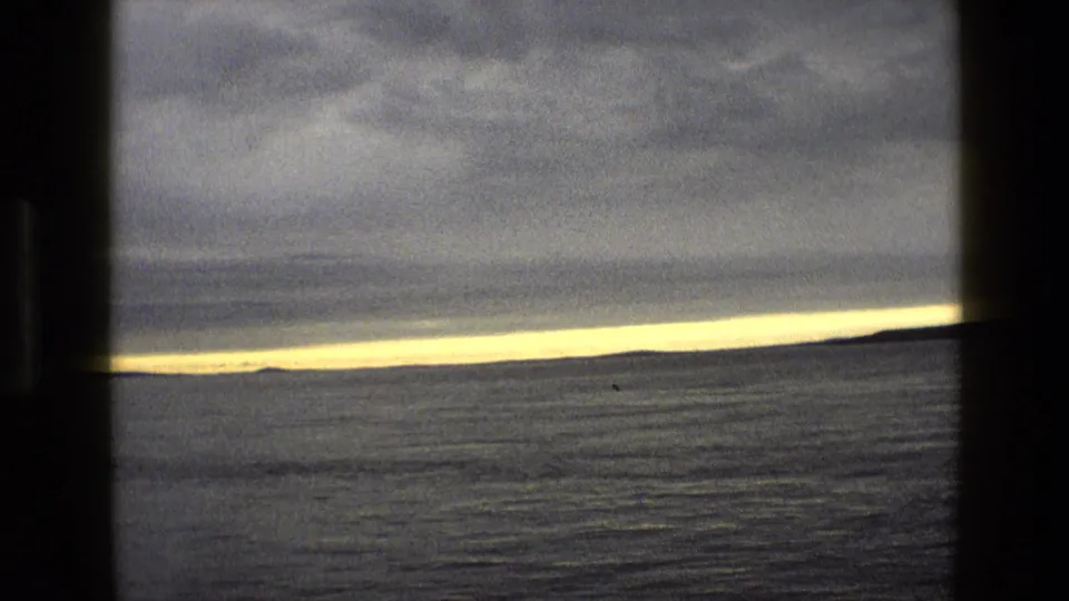 1969 England coastal view across open water, dark cloud cover, and a narrow strip of yellow light near the horizon.
