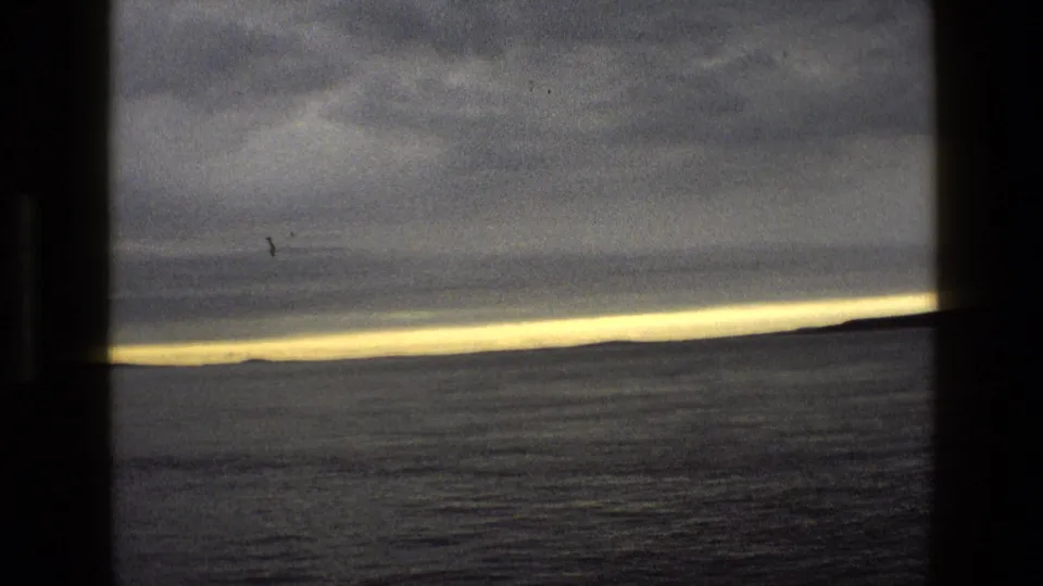 1969 England home-movie frame looking over grey coastal water toward a low horizon beneath dense clouds.