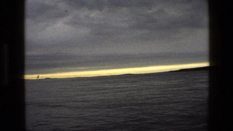 1969 England coastal frame with open water, heavy clouds, and a pale illuminated horizon line across the distance.