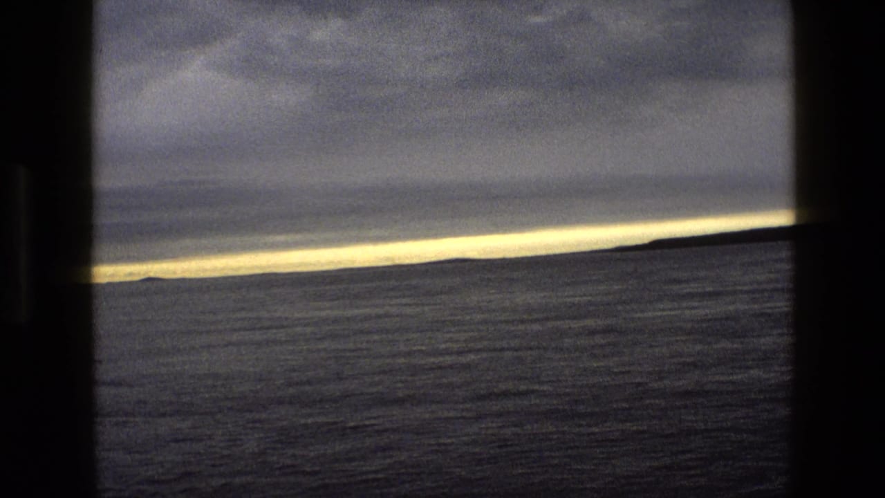1969 England 8mm frame of still water and distant land seen through dark vertical framing as sunset light thins along the horizon.