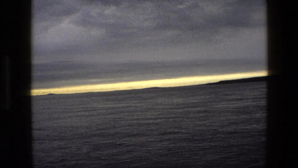 1969 England 8mm frame of still water and distant land seen through dark vertical framing as sunset light thins along the horizon.