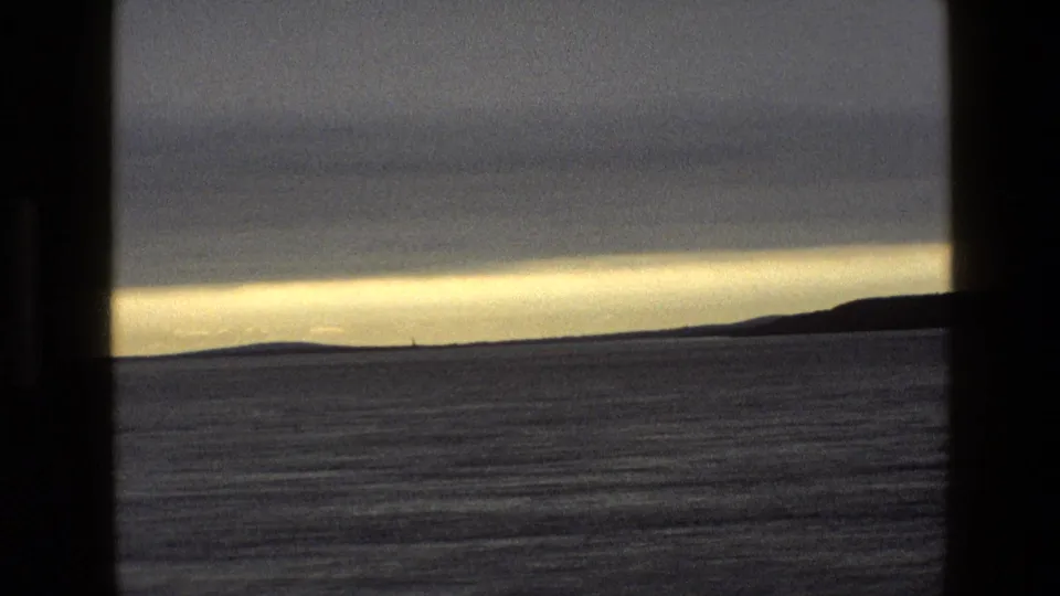 1969 England 8mm frame showing calm sea, low dark land, a small distant marker, and yellow-gray evening light under clouds.