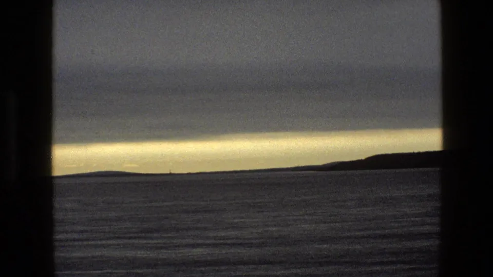 1969 England 8mm frame of quiet open water, a low coastline, and subdued sunset light viewed through dark cabin-like framing.