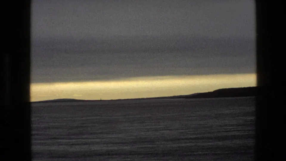 1969 England 8mm home-movie frame showing calm water, distant low hills, heavy clouds, and a thin golden horizon through a window.