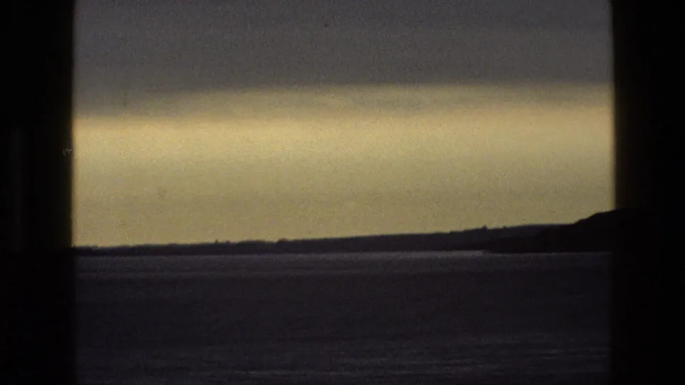 1969 England 8mm home-movie view of water and distant hills under a cloudy twilight sky, seen through dark vertical window bars.