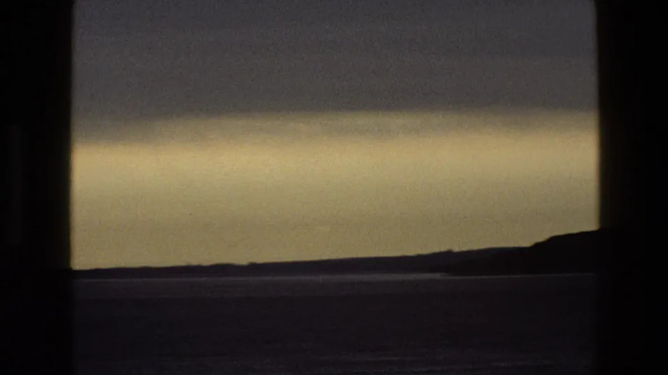 1969 England 8mm frame showing a dim shoreline, calm water, and a pale band of twilight beneath heavy clouds through a window.