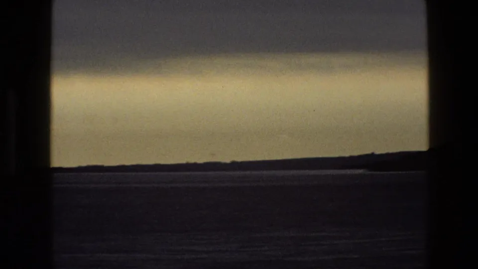 1969 England home-movie frame looking through a dark window at a hazy sky over a low, flat horizon.